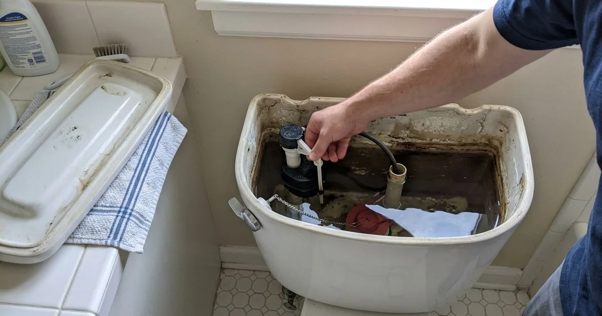 Open toilet tank showing the flapper and fill valve with a hand adjusting the float